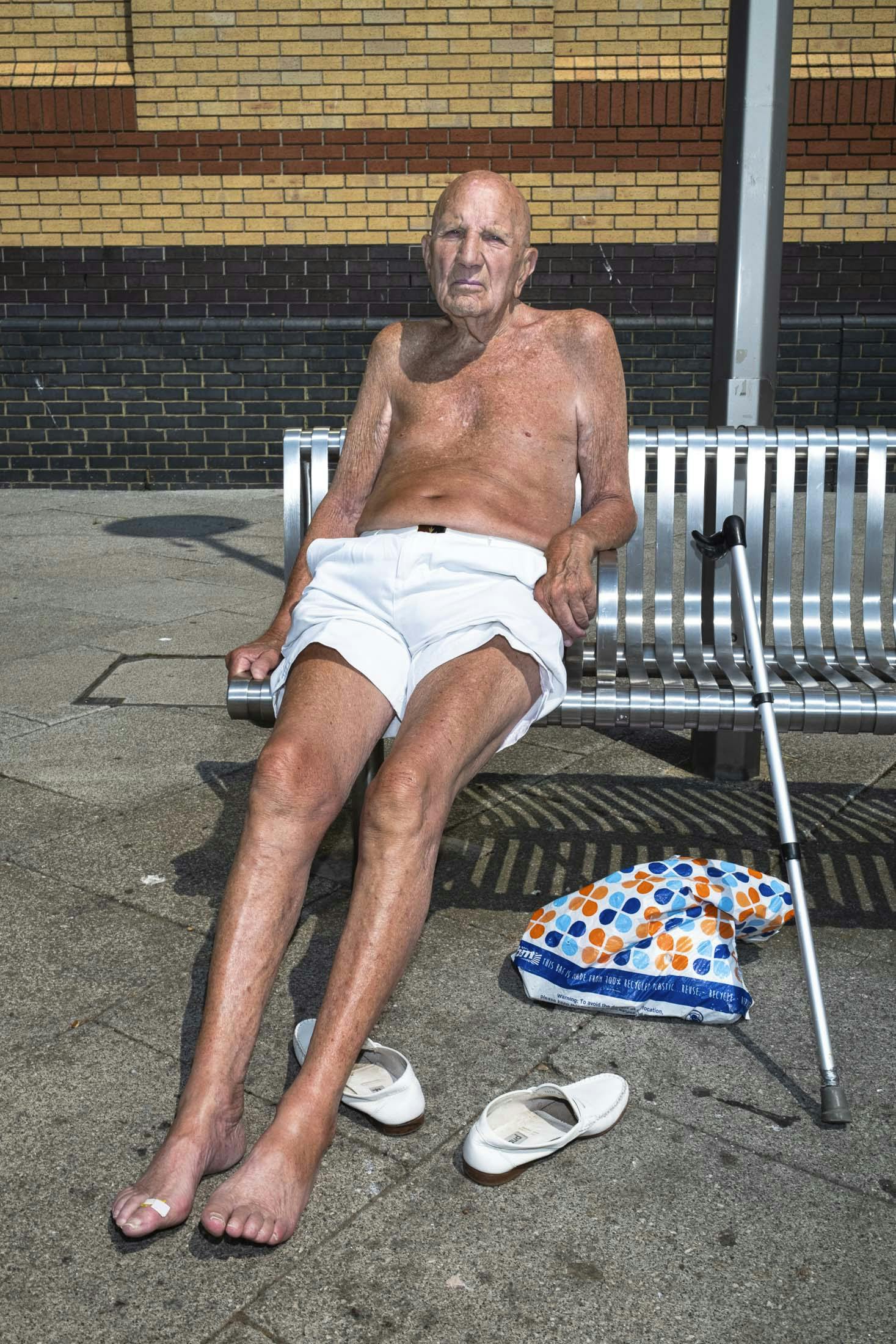 Shirtless old man in white shorts sun bathing on bench in Southend England