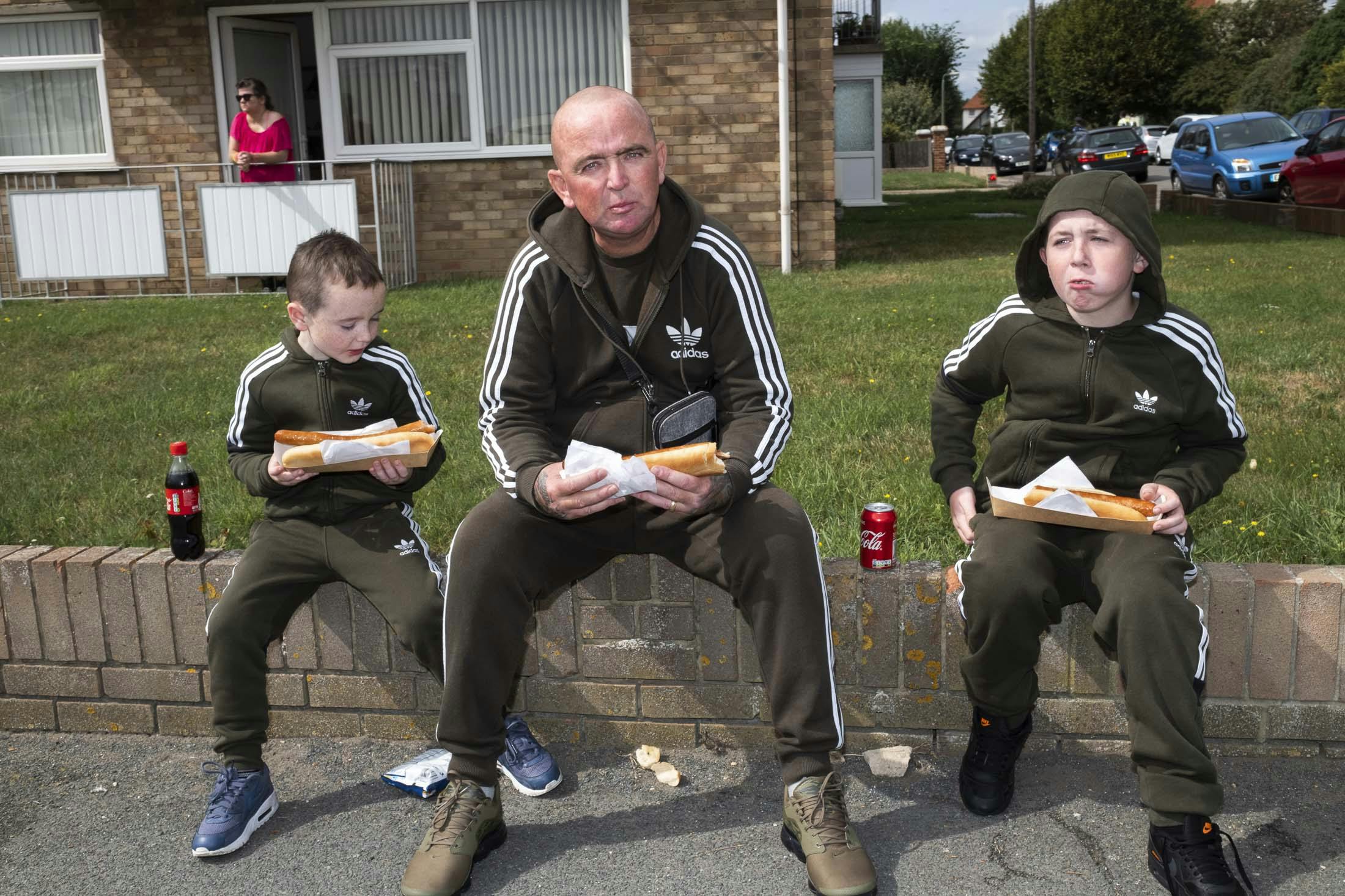Dad and sons wearing matching green adidas tracksuis eating hotdogs and drinking coke