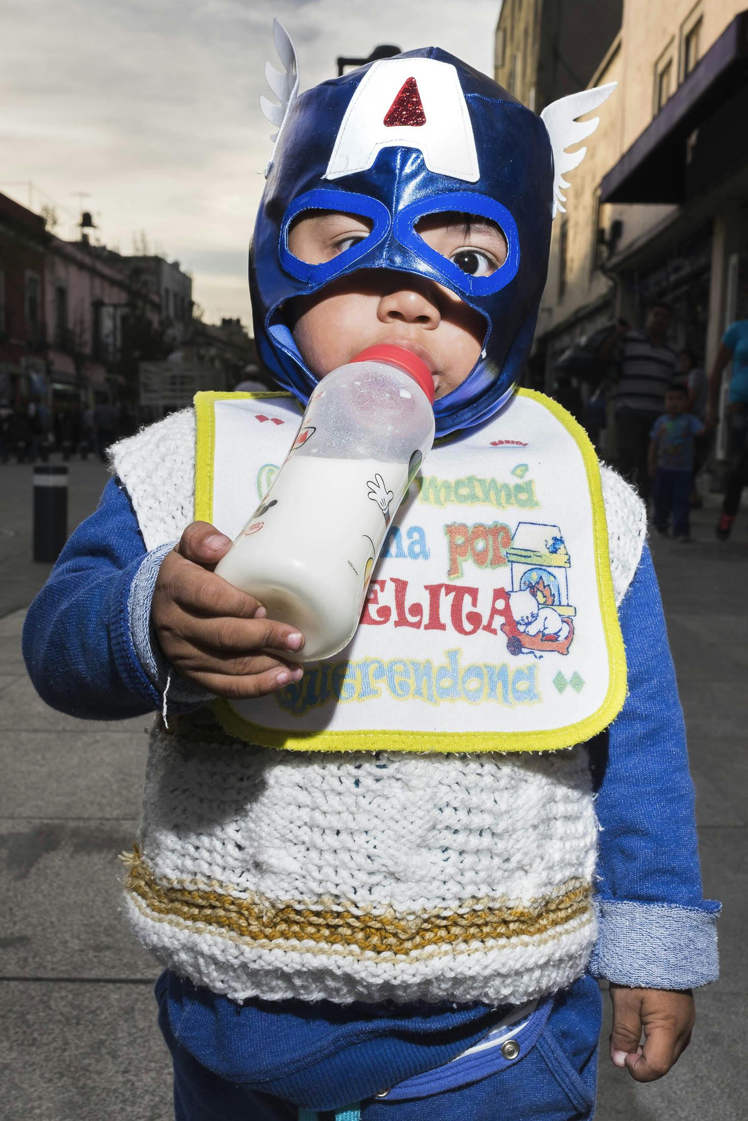 Boy dressed as Captain America walking down a Mexican street drinking a bottle of milk